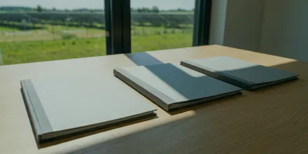 Drei geschlossene Mappen nebeneinander auf einem Holztisch am Fenster, draußen eine sonnige Wiese