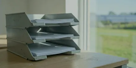 Dreistöckige Ablage aus Metall mit Papierstapeln auf einem Schreibtisch, draußen eine Freiflächenanlage