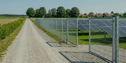 Schotterweg entlang des Zauns einer Freiflächen-Solaranlage, im Hintergrund Bäume und Wohnhäuser