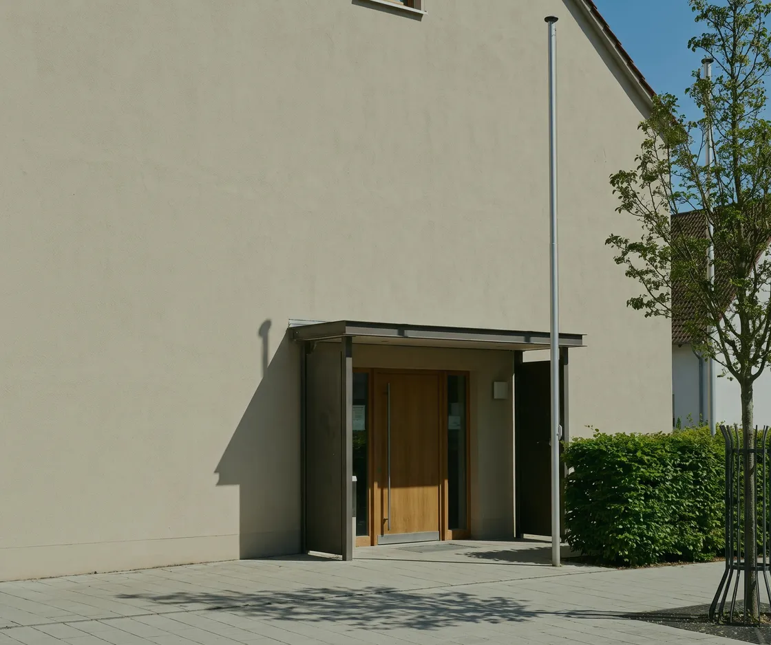 Schlichtes Verwaltungsgebäude mit Vordach und Holztür, davor eine geschnittene Hecke und ein junger Baum