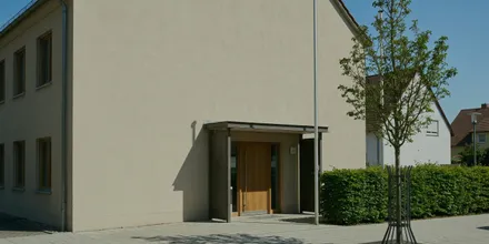 Schlichtes Verwaltungsgebäude mit Vordach und Holztür, davor eine geschnittene Hecke und ein junger Baum