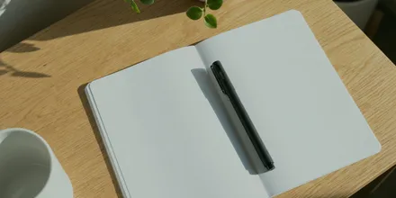 Aufgeschlagenes leeres Notizbuch mit Stift auf einem Holztisch, daneben eine Tasse und eine Pflanze