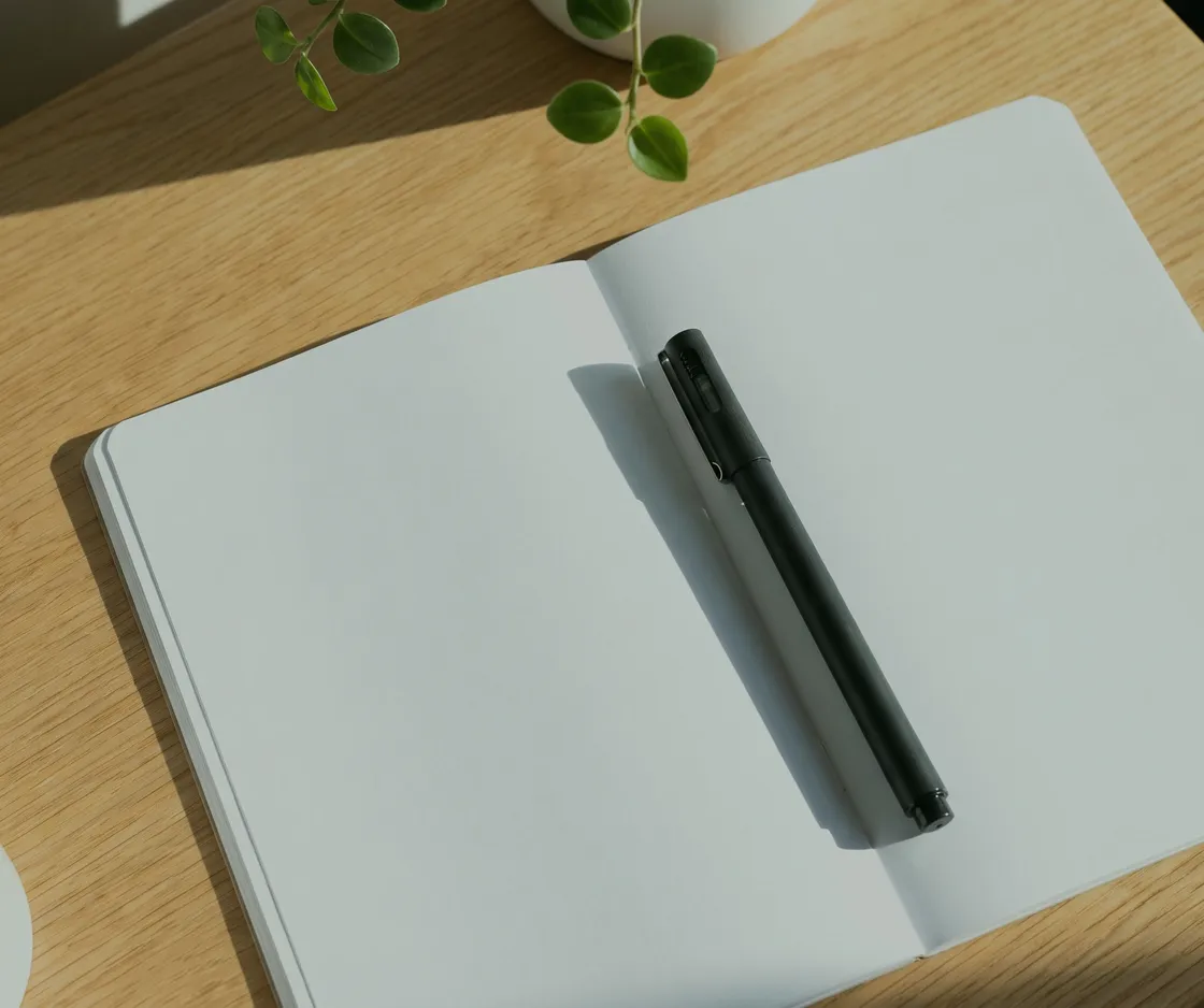 Aufgeschlagenes leeres Notizbuch mit Stift auf einem Holztisch, daneben eine Tasse und eine Pflanze