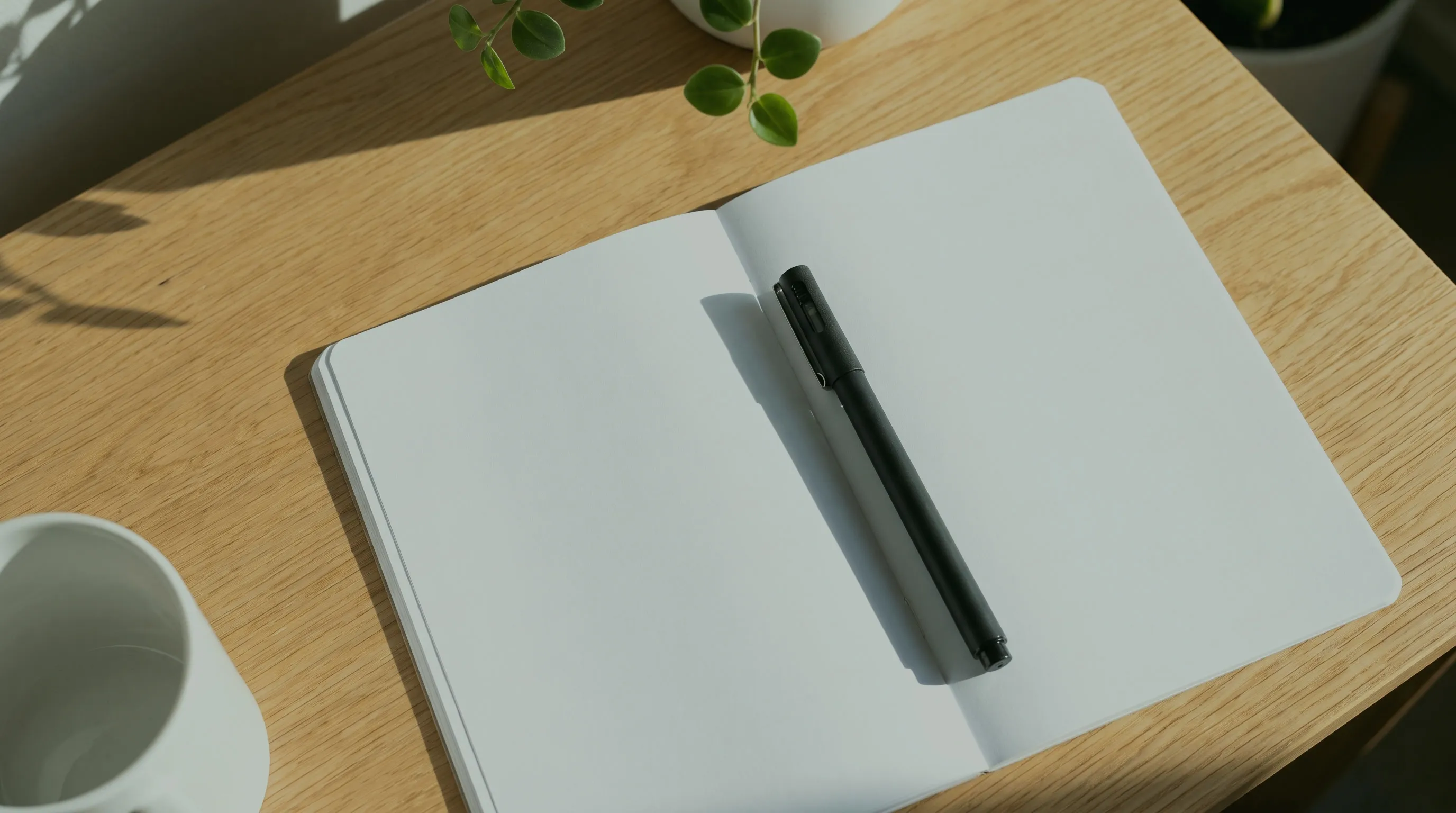 Aufgeschlagenes leeres Notizbuch mit Stift auf einem Holztisch, daneben eine Tasse und eine Pflanze