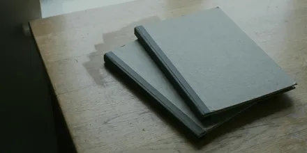 Zwei graue Hefte mit festem Einband, leicht versetzt übereinander auf einem alten Holztisch