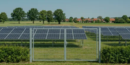 Geschlossenes Tor im Zaun einer Freiflächen-Solaranlage, dahinter Modulreihen und ein Dorf